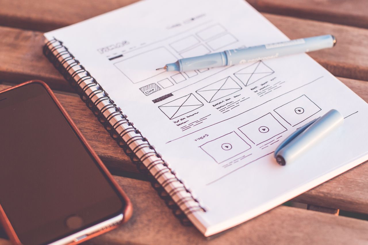 Vue rapprochée d’un carnet avec des esquisses de wireframe et un smartphone posé sur un bureau en bois.